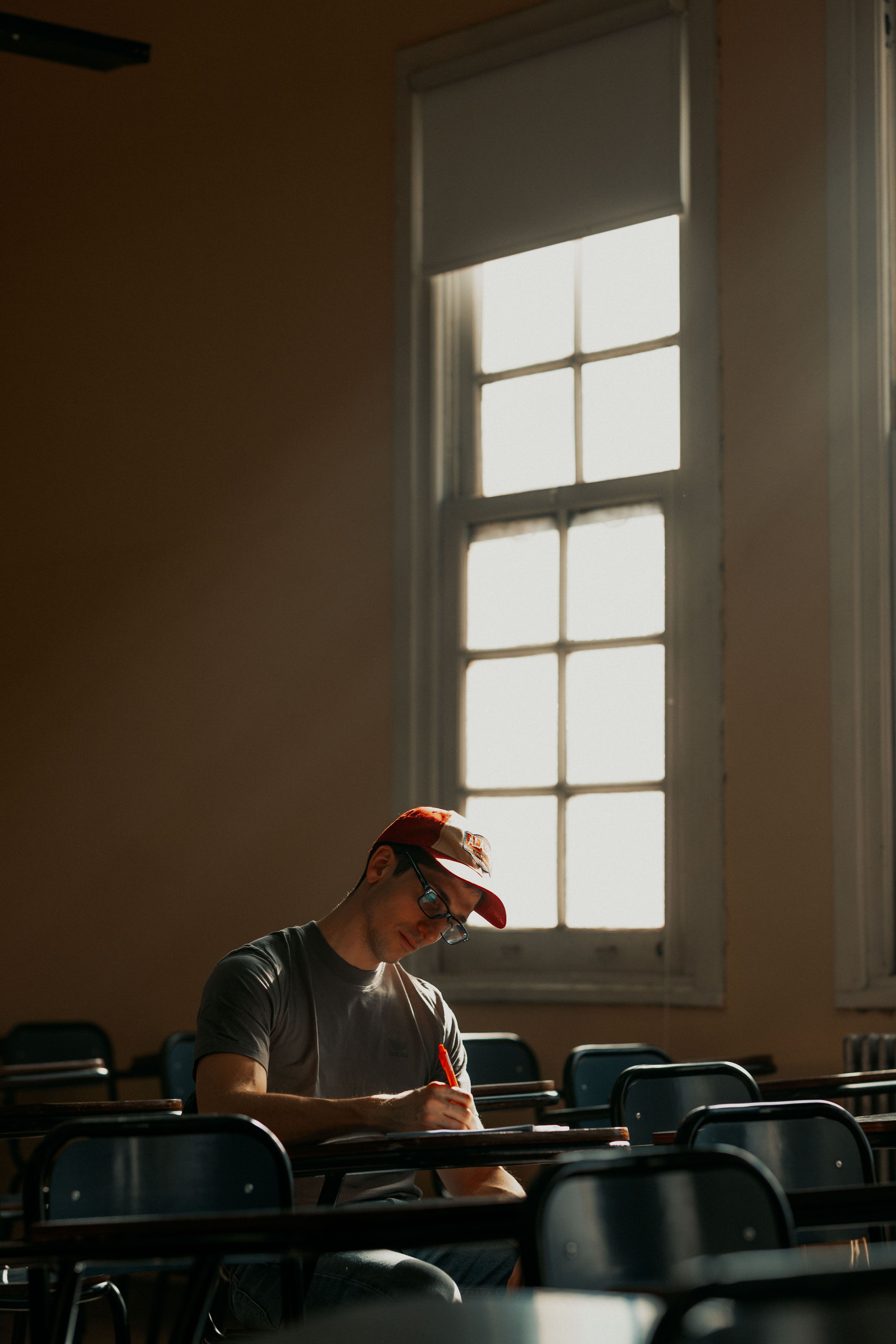A student wearing a cap studies diligently in a sunlit classroom.