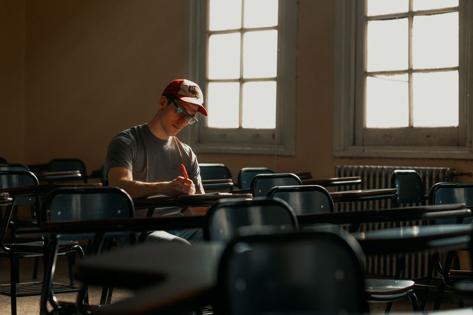 A young man in a cap studies alone in an empty classroom filled with desks. A young man in a cap studies alone in an empty classroom filled with desks.
