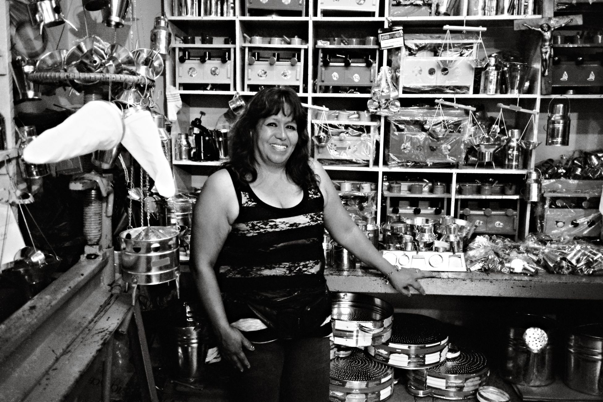 A female shop owner smiling amidst metal goods in a Quito market.