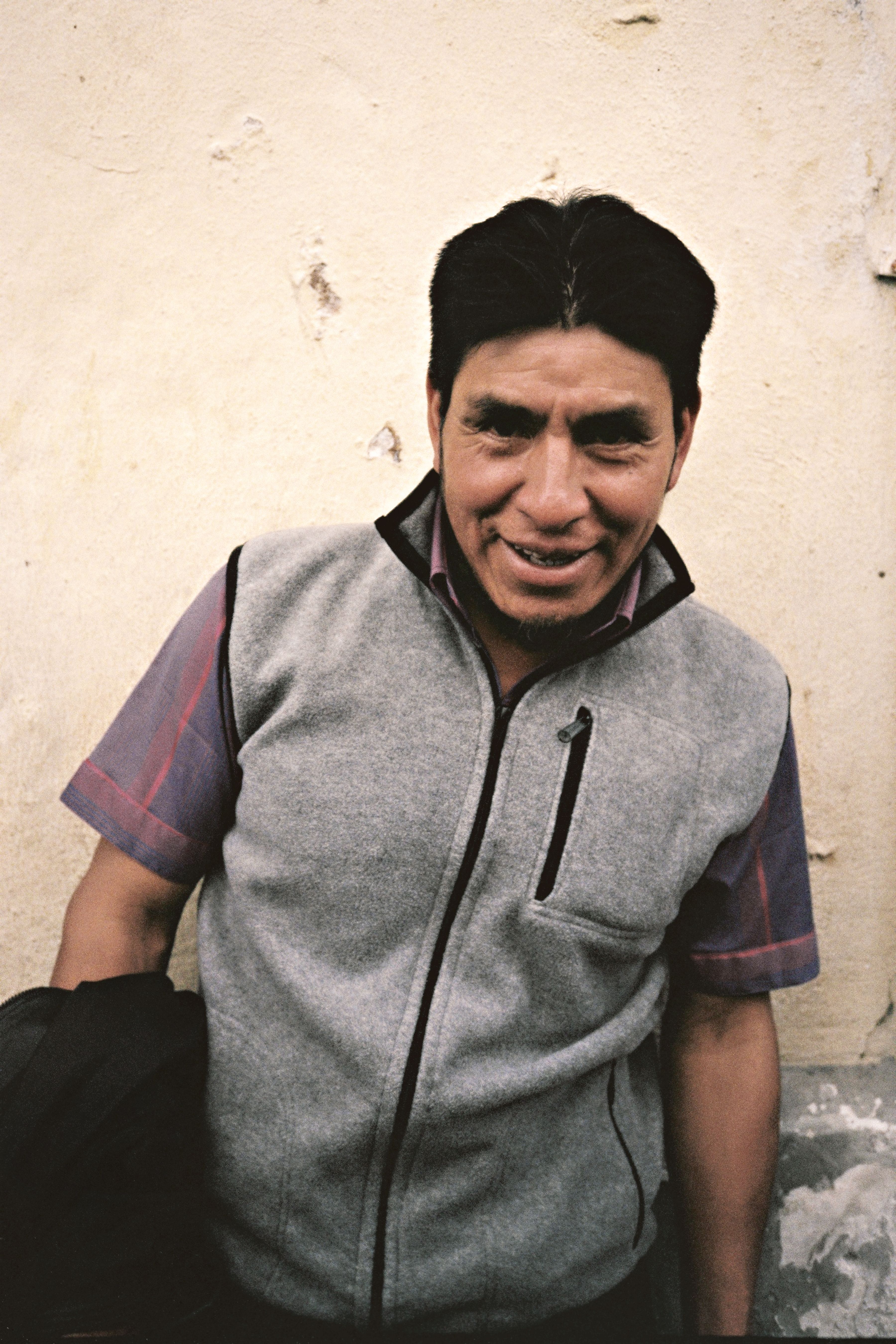 A man in casual wear smiling at the camera near a rustic wall in Quito, Ecuador.