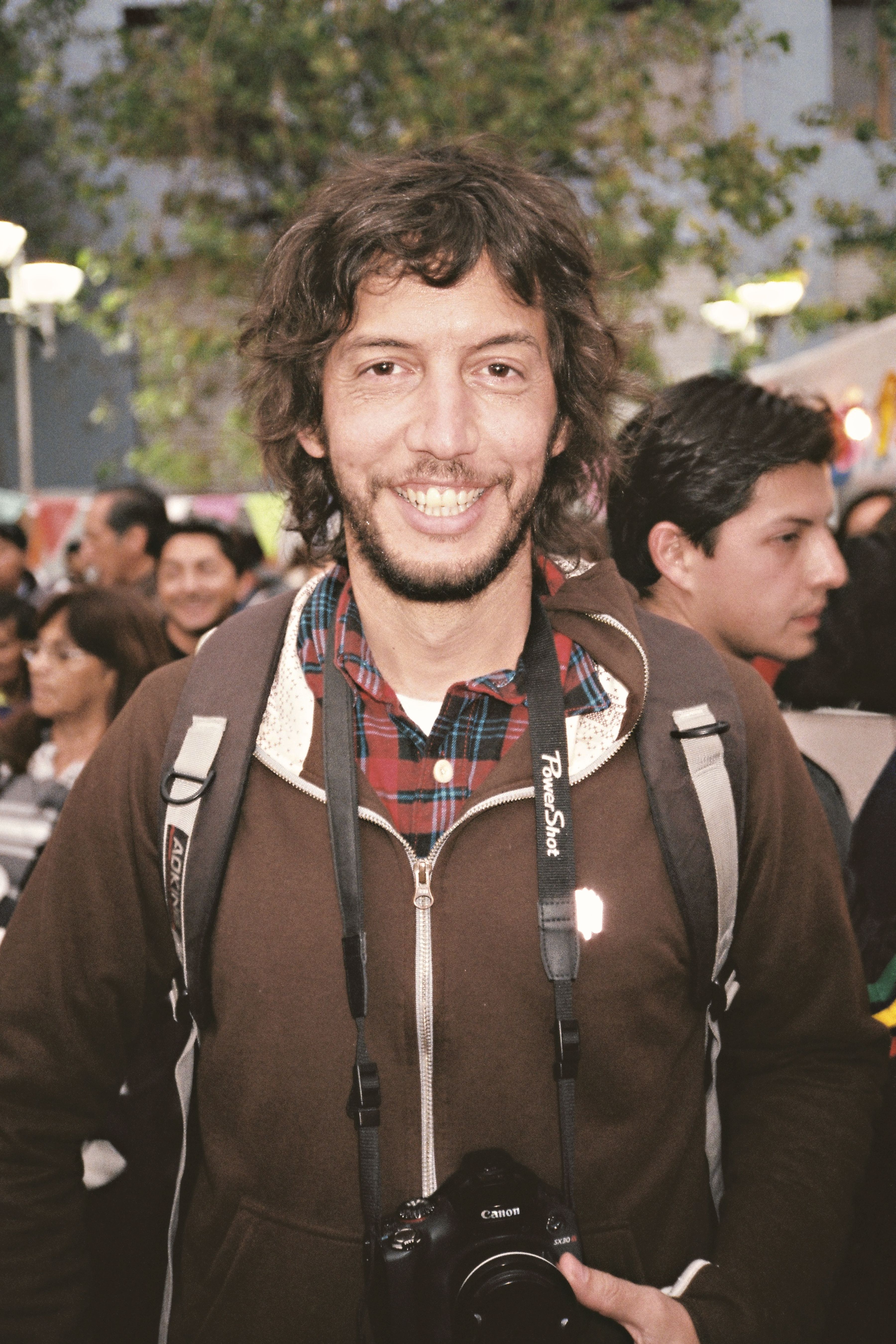 A smiling photographer holding a camera amidst a lively crowd outdoors in Quito, Ecuador.