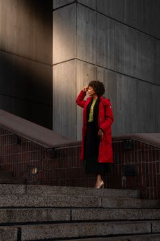 Confident woman in vibrant red coat poses on city steps against modern architecture.