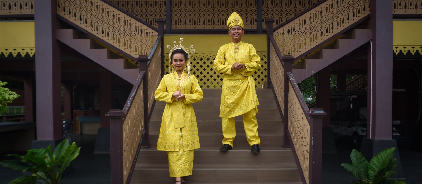Couple in bright yellow Malay traditional clothing descending a staircase indoors.