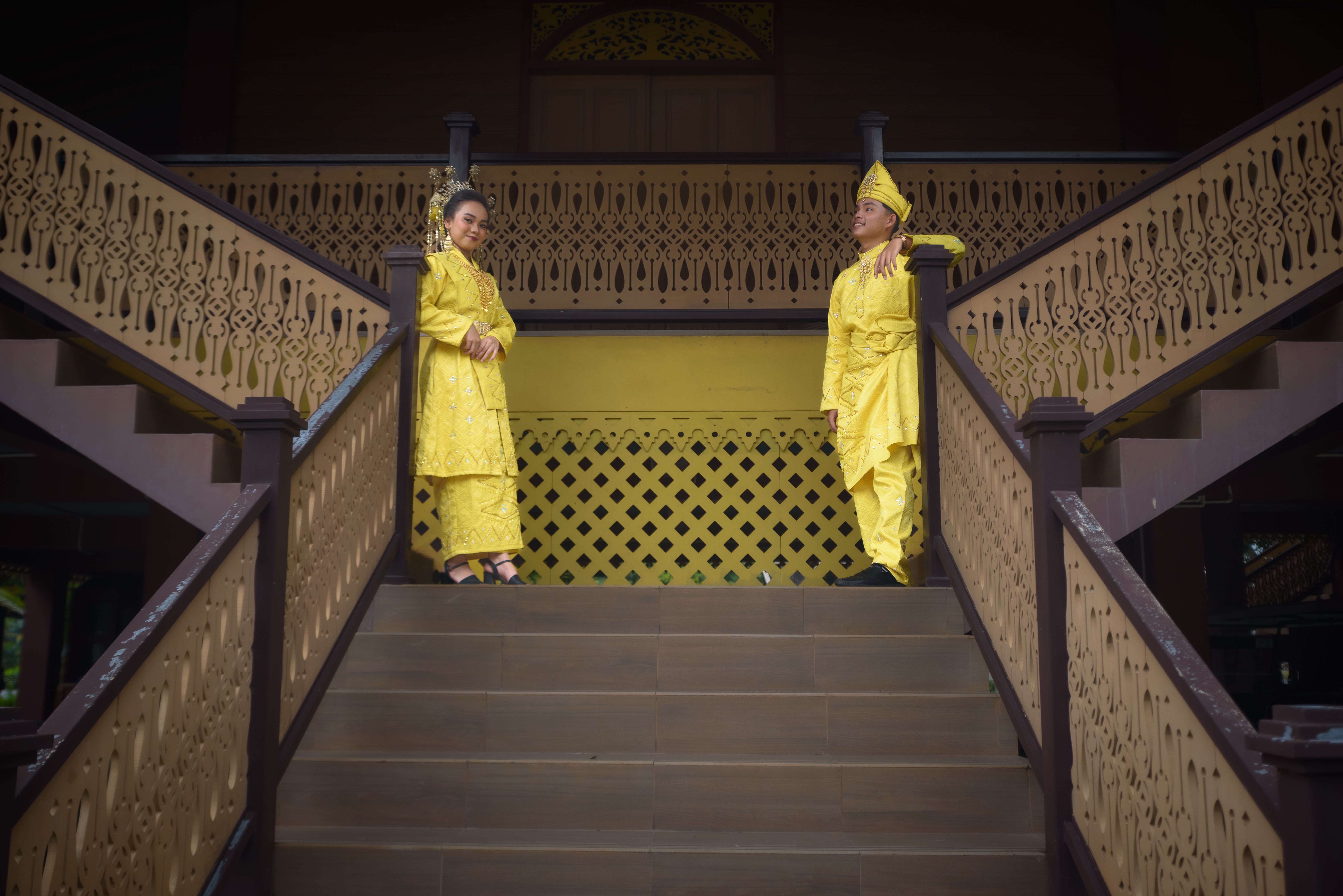 Two adults in vibrant traditional yellow attire on intricately carved wooden stairs.