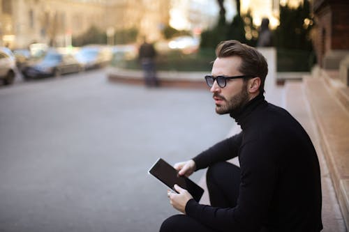 Side view of trendy serious bearded male in glasses and black casual clothes with notebook looking away and thinking while sitting on exterior stairs at entrance of modern building in downtown
