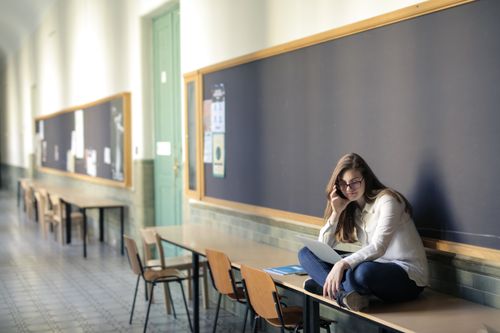Free A woman sits on a desk in a university hallway, studying on a laptop. Stock Photo