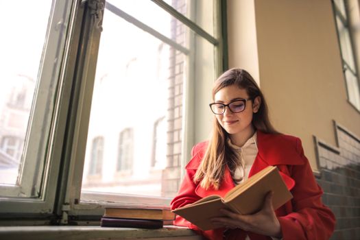 A woman in a red coat reads a book by the window, smiling in natural light.. woman, coat, reads
