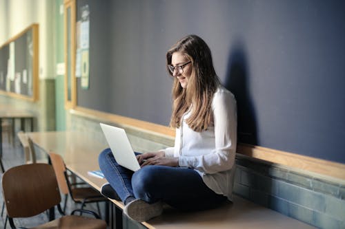 Woman in White Long Sleeve Shirt and Blue Denim Jeans Sitting on Table While Using Macbook