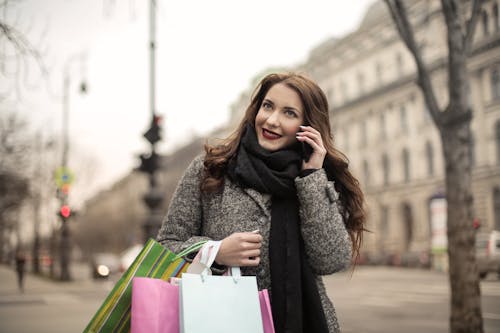 Happy adult woman in warm coat and scarf with shopping bags smiling away while standing on street and talking on phone against blurred urban environment in cool overcast weather