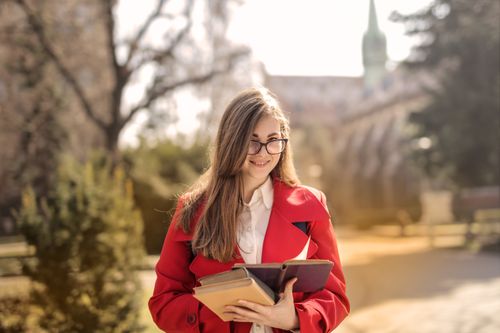 Free Smiling young woman in red coat holding books outdoors with a soft focus background. Stock Photo