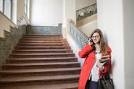 一位穿着红色外套的年轻女子在室内微笑着，一边用智能手机，一边拿着书