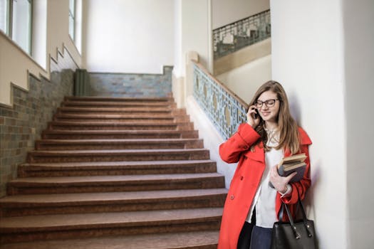 一位穿着红色外套的年轻女子在室内微笑着，一边用智能手机，一边拿着书