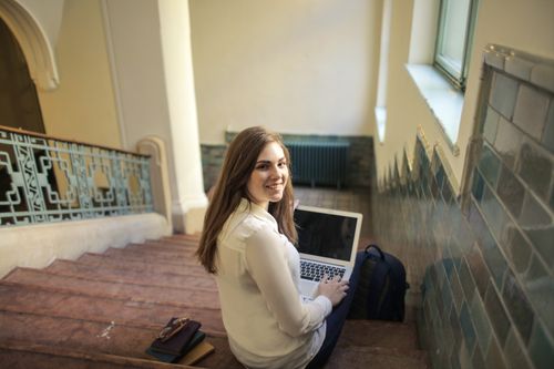 Free A young woman sits on a staircase using a laptop indoors, smiling back at the camera. Stock Photo