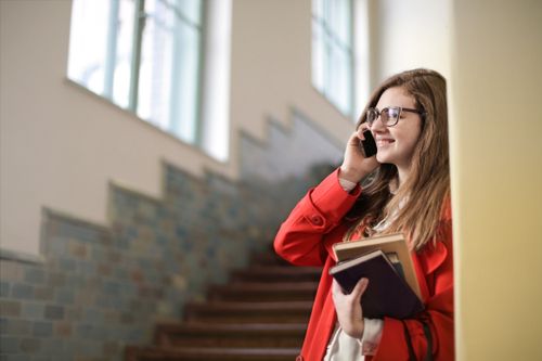 Free A woman in a red coat talks on her smartphone, smiling and holding books, indoors. Stock Photo