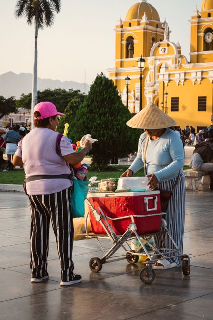 Two street vendors interact in front of a vibrant yellow church in Trujillo's historic plaza at dusk.