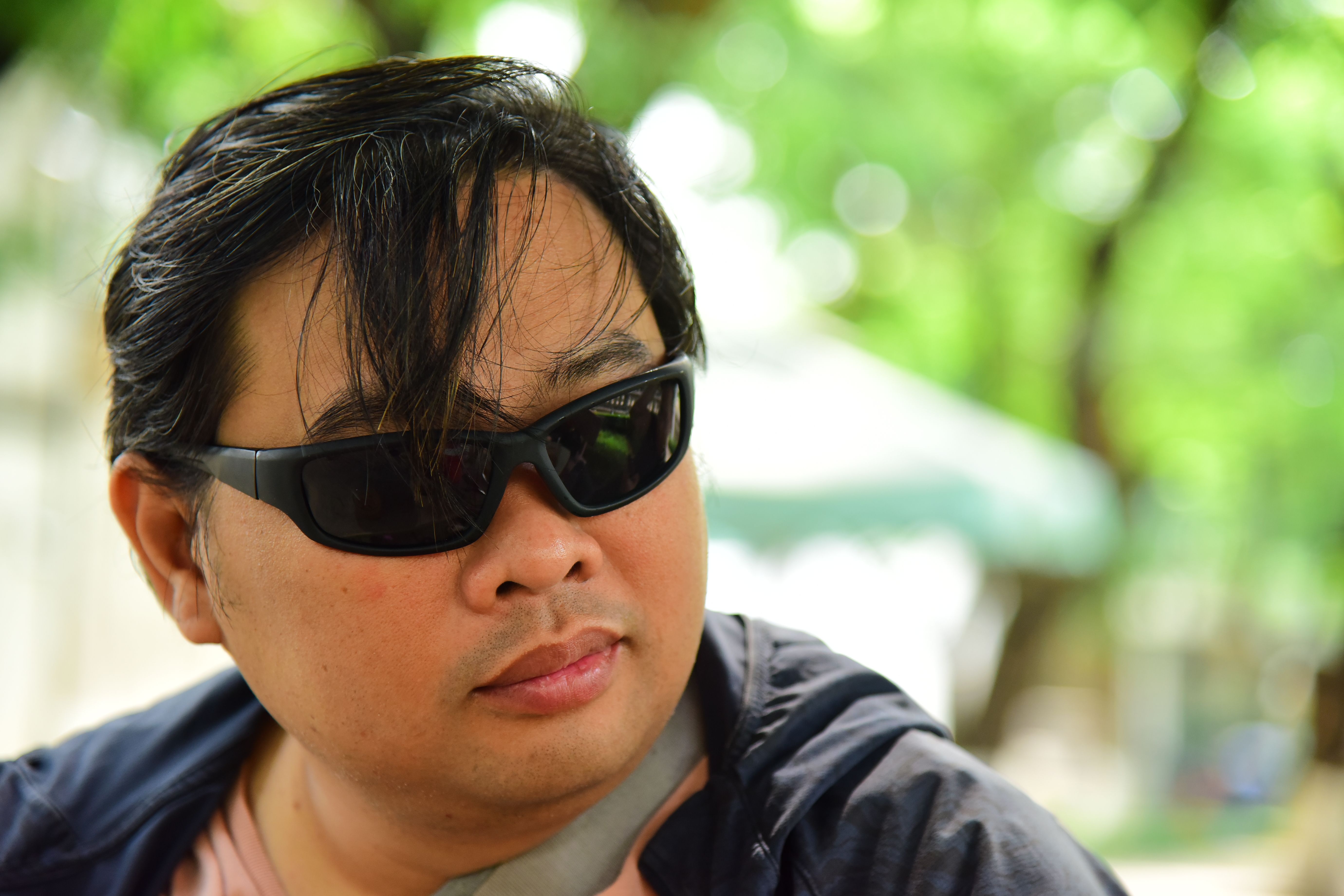 Close-up portrait of an Asian man wearing sunglasses outdoors in a sunny park.