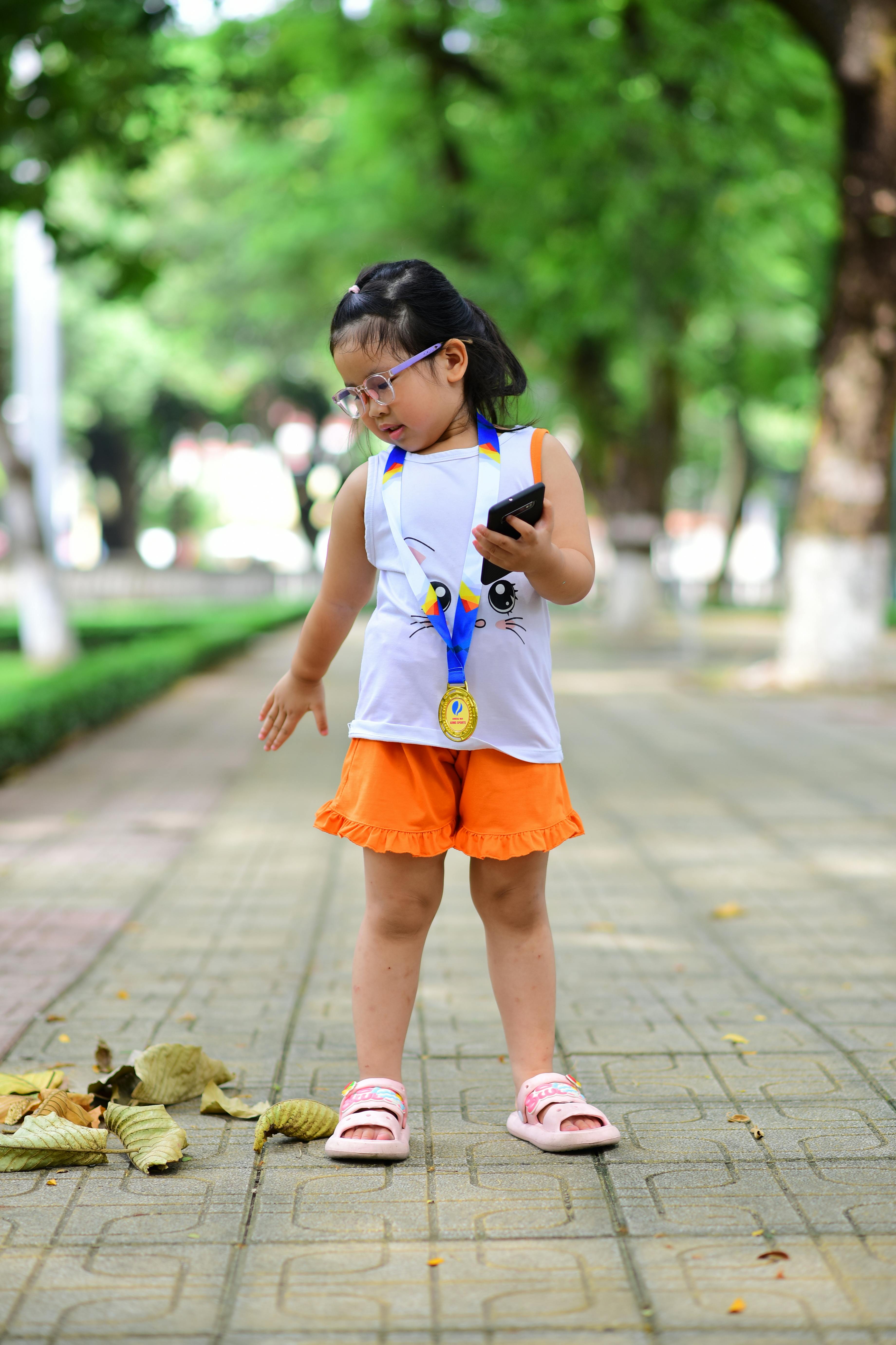 A young girl wearing a medal and holding a smartphone stands on a brick sidewalk outdoors.