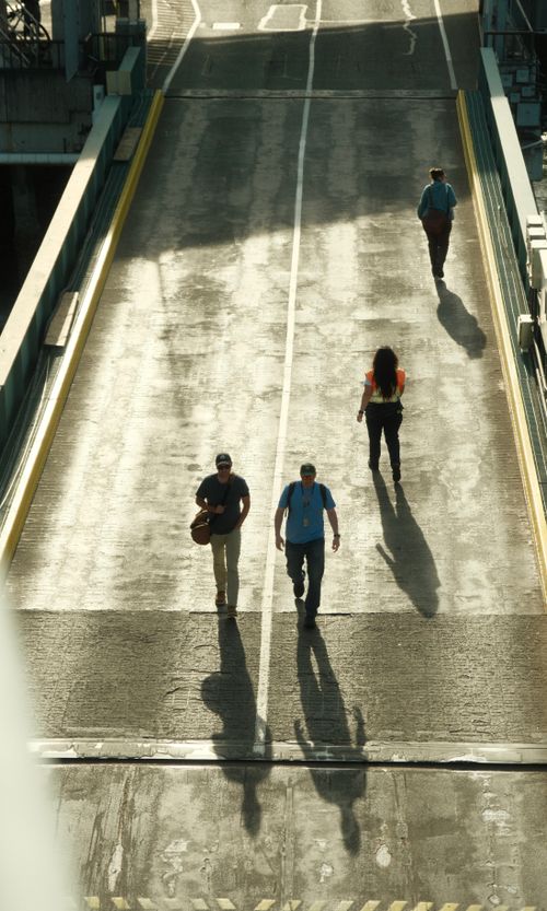 Free Four individuals walking on a sunlit bridge in an urban setting, casting long shadows on the asphalt. Stock Photo