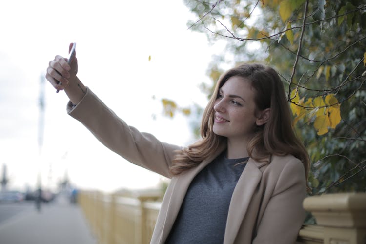 Young Woman In Coat Taking Selfie With Smartphone On Street In Overcast Weather