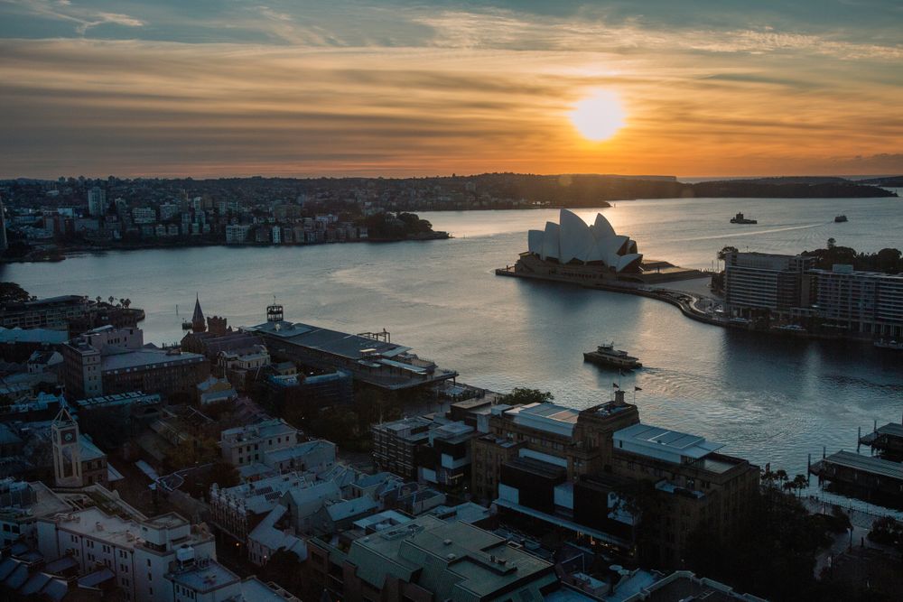 Vista aérea de Sydney al ocaso con la Opera House recortada contra el sol bajo y la bahía dorada