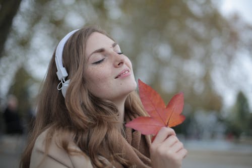 Junge Zufriedene Frau In Den Kopfhörern Mit Frischem Rotem Blatt, Das Musik Mit Vergnügen Beim Faulenzen Im Herbstpark Hört