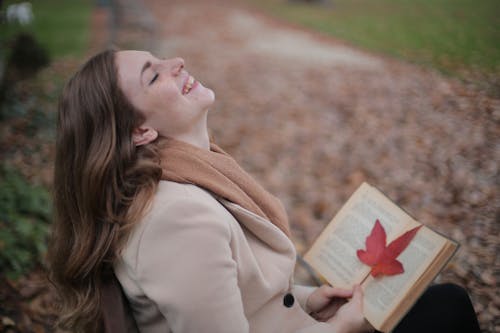 Веселая молодая женщина с красным листом наслаждается жизнью и погодой, читая книгу в осеннем парке