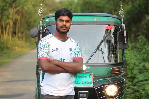 Free Man standing confidently in front of a green rickshaw on a peaceful rural road. Stock Photo