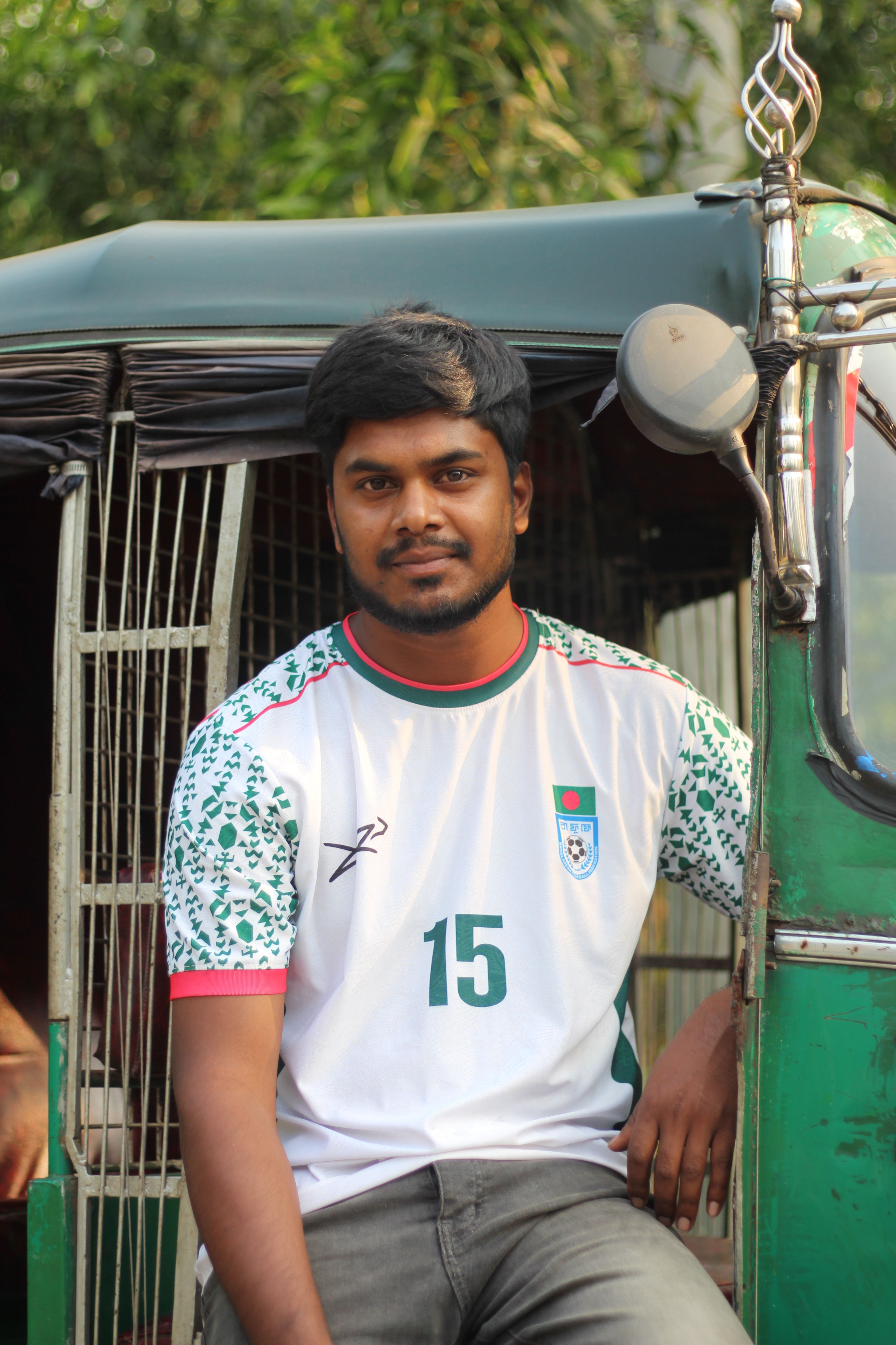 A young man sits in a rickshaw wearing a sports jersey with a relaxed smile on a sunny day.