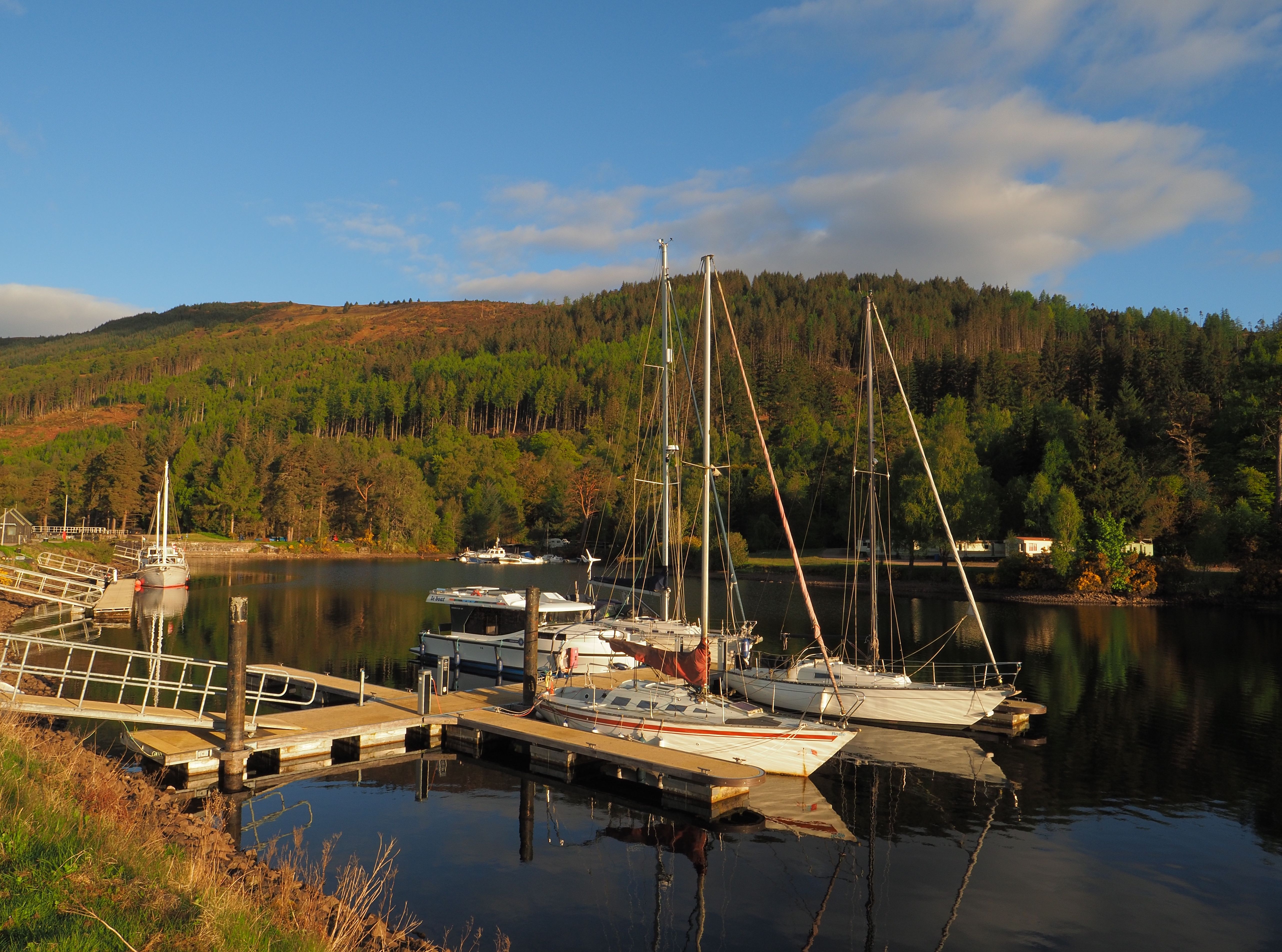 Peaceful marina in Scotland with sailboats docked, surrounded by lush hills at sunrise.