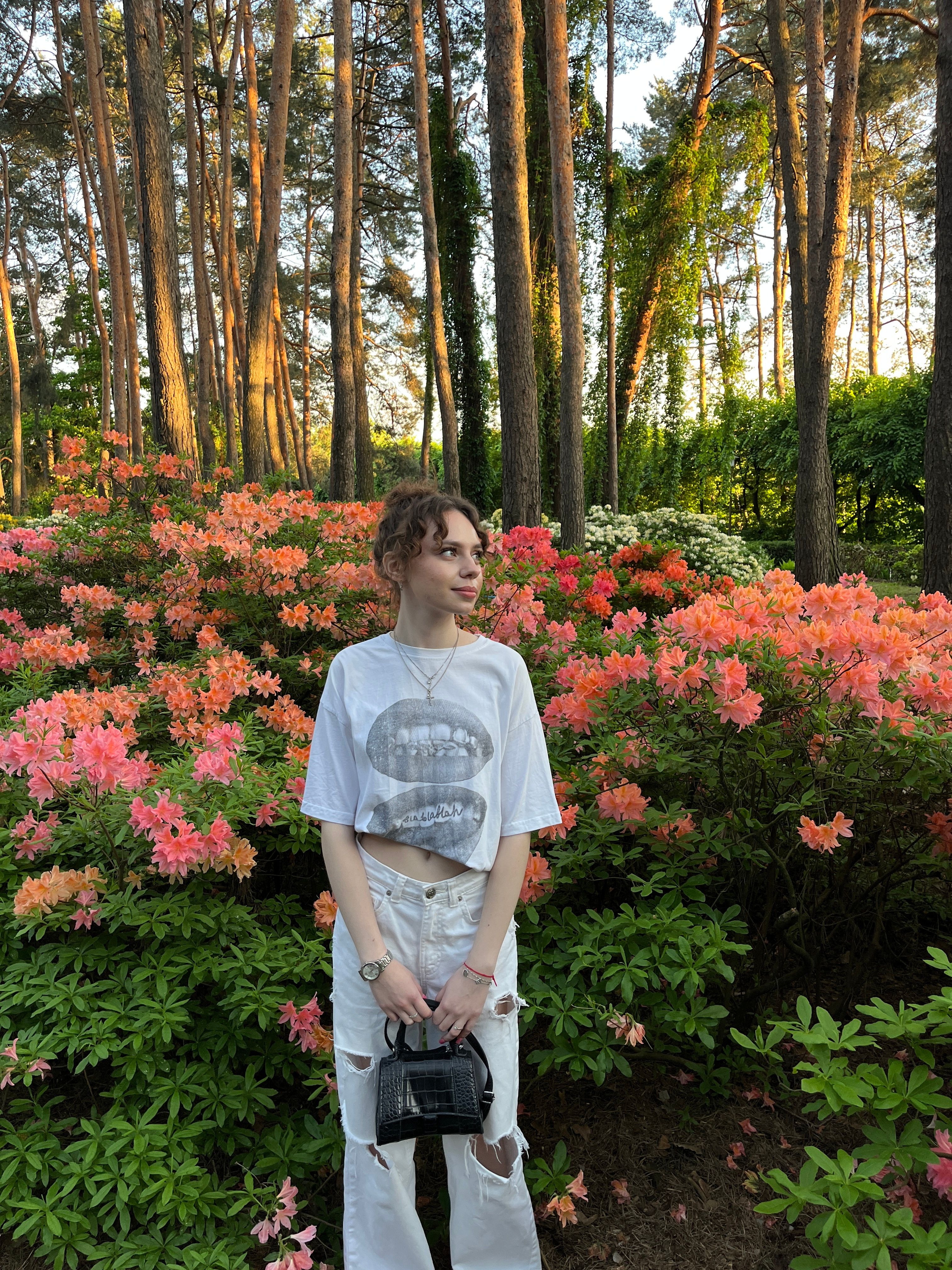 A young woman stands among colorful azaleas in a sunlit forest setting, exemplifying natural beauty.