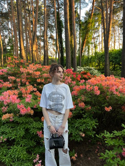 A young woman stands among colorful azaleas in a sunlit forest setting, exemplifying natural beauty.