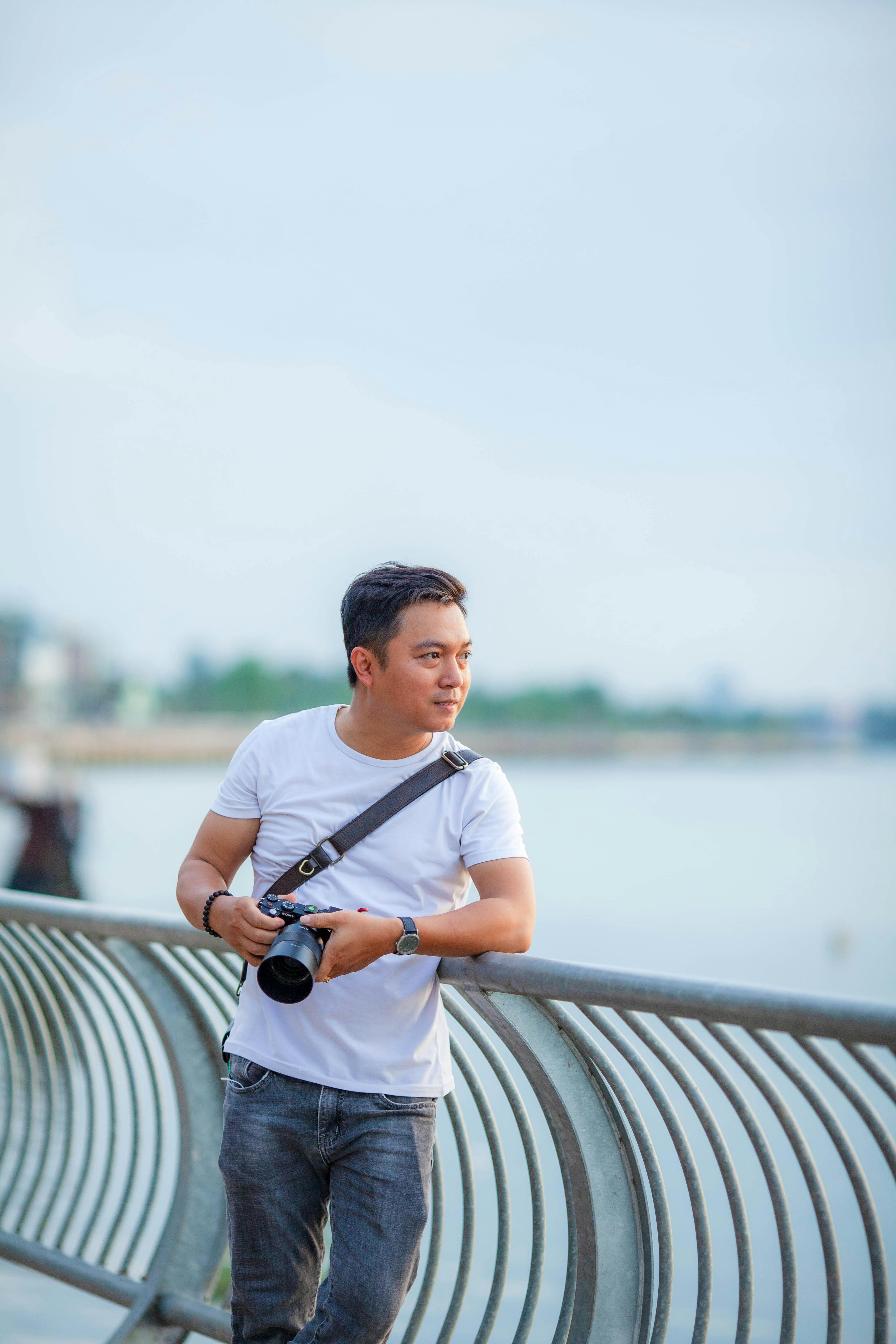 A photographer with a camera leans on a railing by the water on a sunny day.