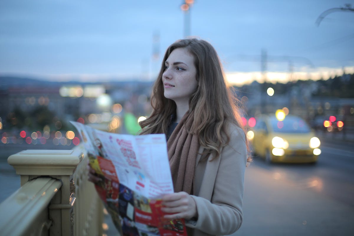 Female traveler in warm clothes standing on street with map and looking away at dusk