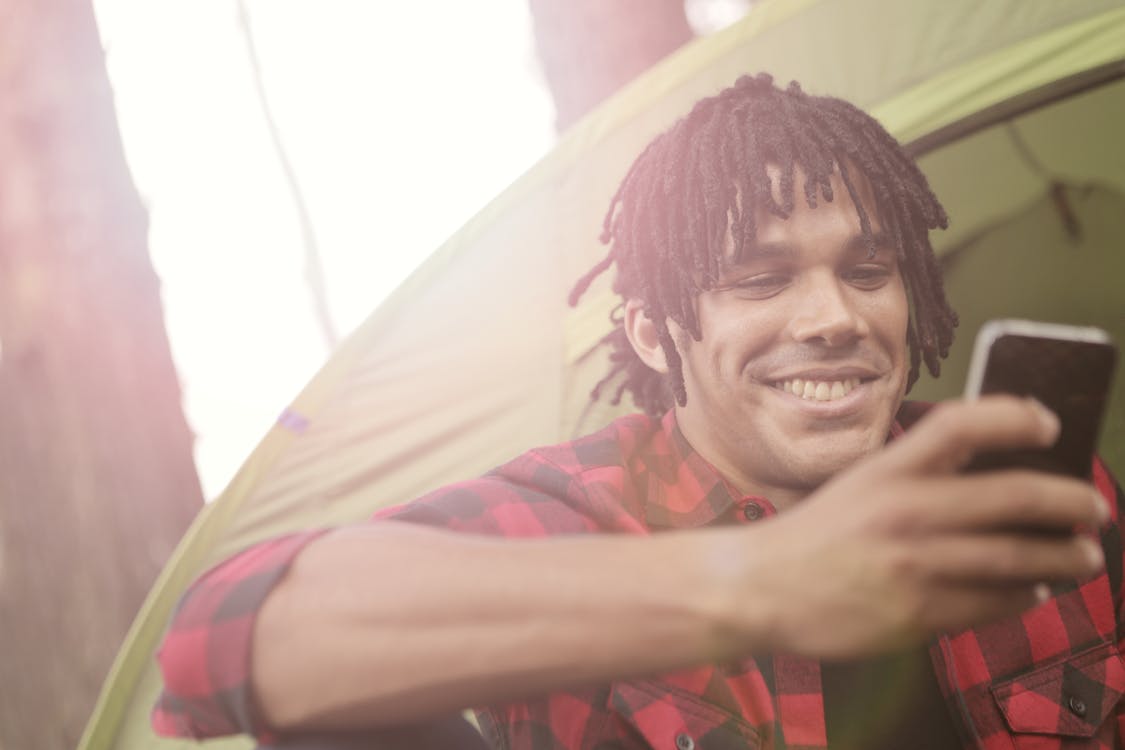 Young casually dressed man smiling while sitting near tent and surfing smartphone in back lit