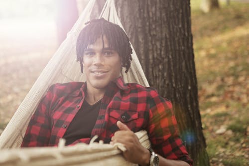 Cheerful man with dreadlocks lying in hammock while smiling and looking at camera
