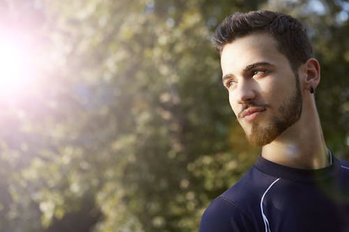 Handsome guy in sportswear and with earring standing in park in sunny summer day and looking away