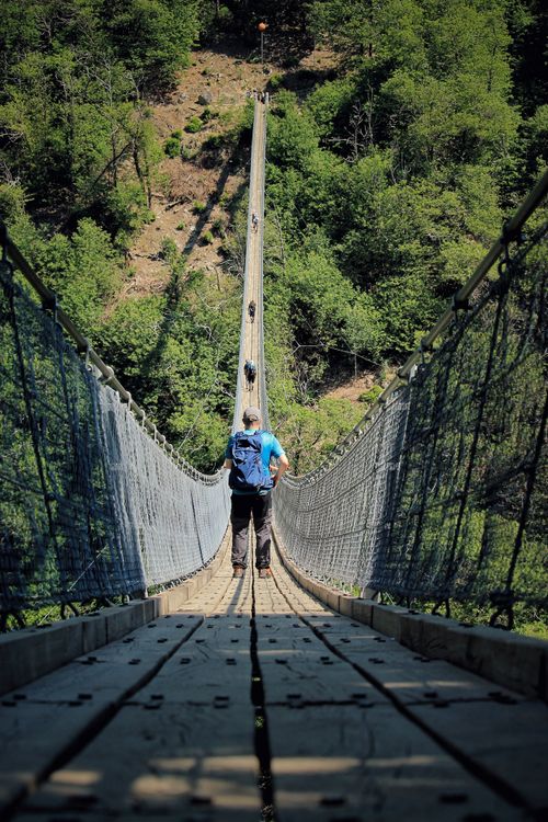 Free A lone hiker crosses a long suspension bridge amidst lush greenery in Ticino, Switzerland. Stock Photo