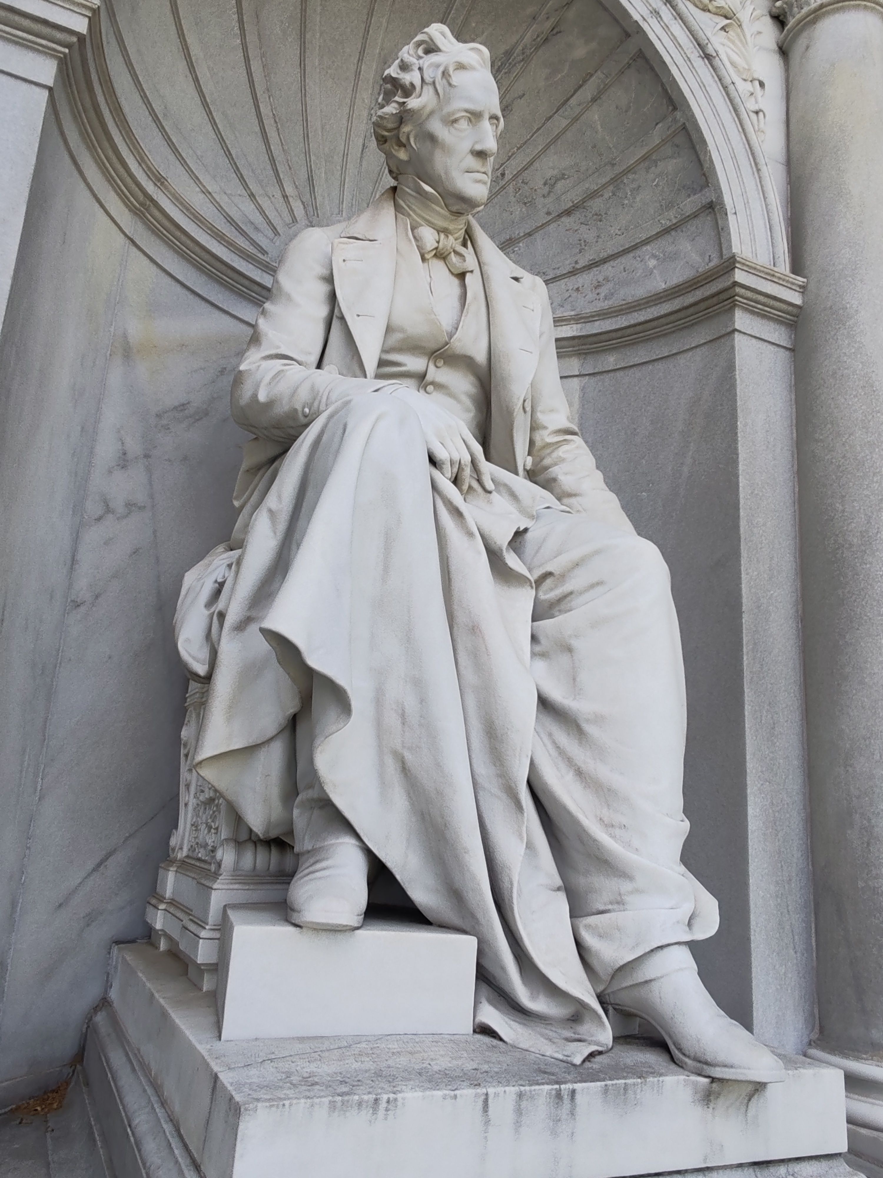 Elegant white marble statue of a historic figure in classical attire against architectural backdrop.