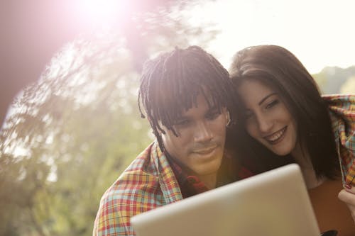 Cheerful man with dreadlocks and woman smiling and browsing tablet while spending time together in park in summer day