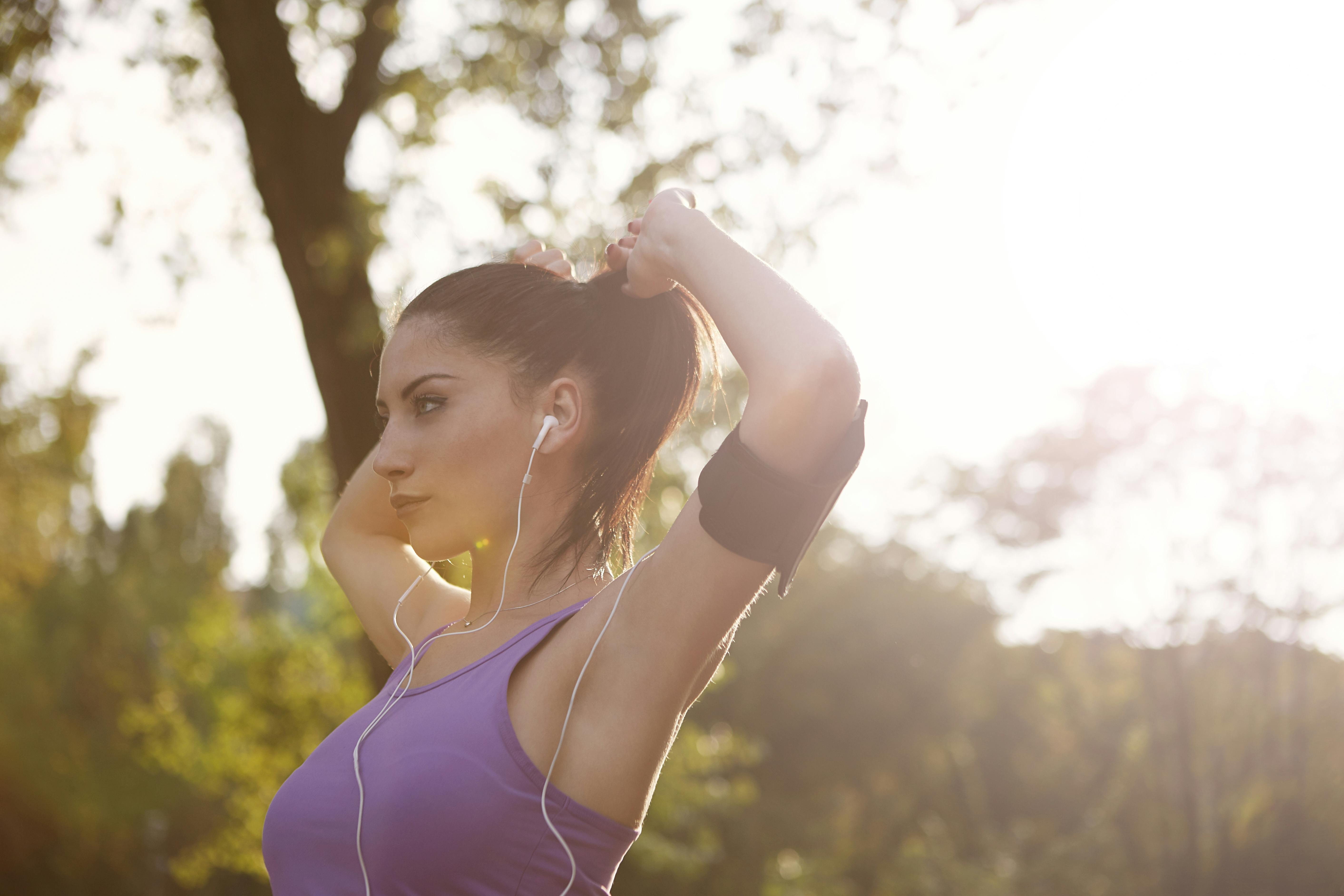 A focused woman adjusts her hair while listening to music during an outdoor workout in a sunlit park.
