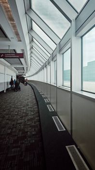 Long airport corridor with travelers and luggage, bright daylight through glass windows.
