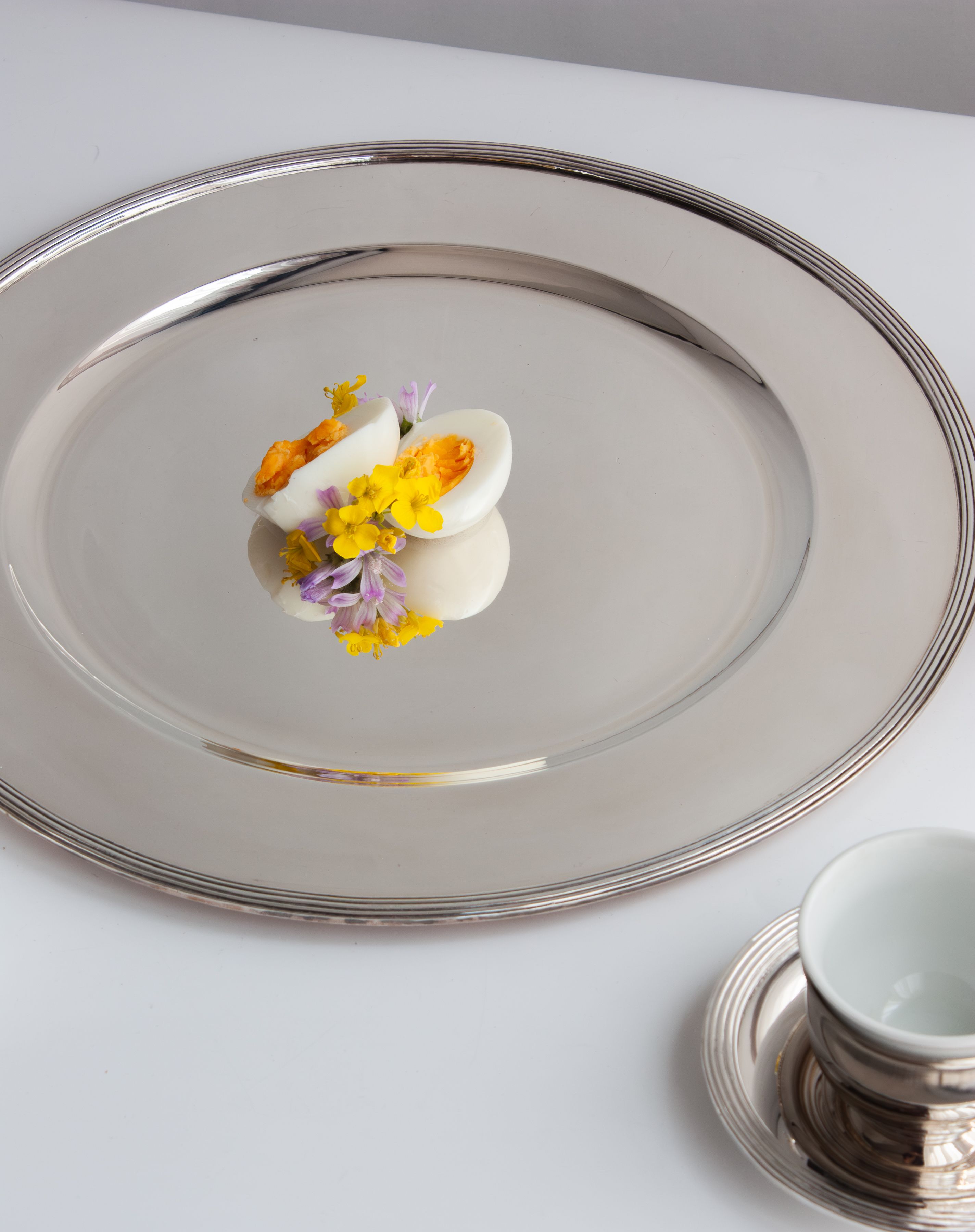 Sophisticated display of a half-boiled egg garnished with yellow and purple flowers on a silver platter. Minimalist and artistic.