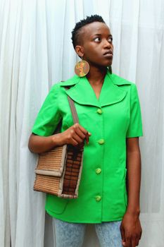 Stylish young woman in green attire with wicker bag and bold earrings against a white curtain.