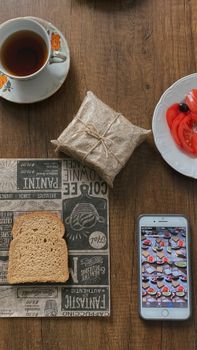 Top-down view of a rustic breakfast setting with tea, bread, tomatoes, and a smartphone on a wooden table.