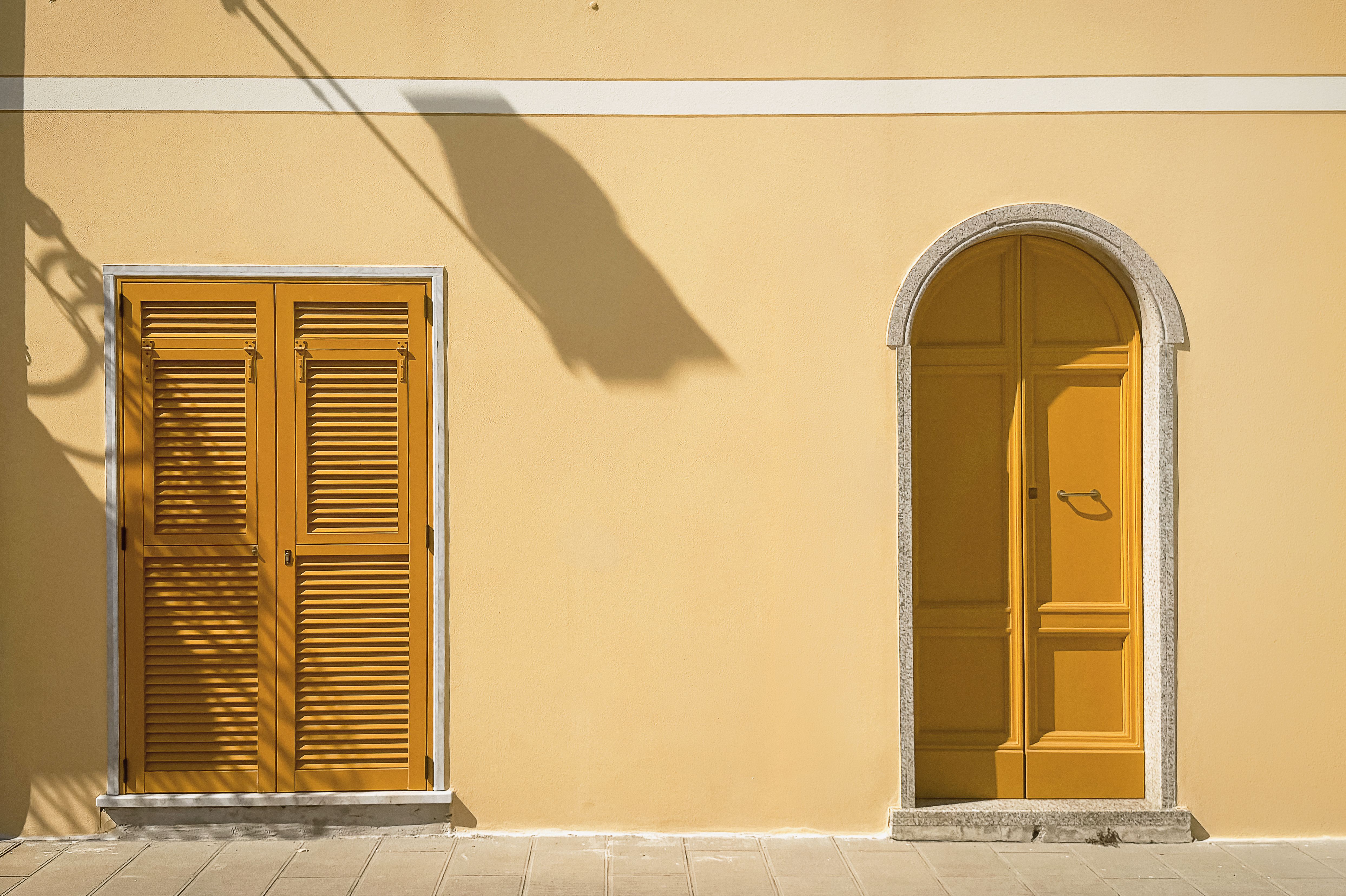 Yellow arched door and shutters on a pastel facade, casting shadows.