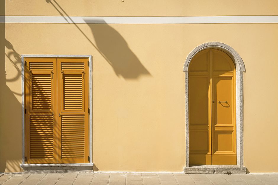 Yellow arched door and shutters on a pastel facade, casting shadows.