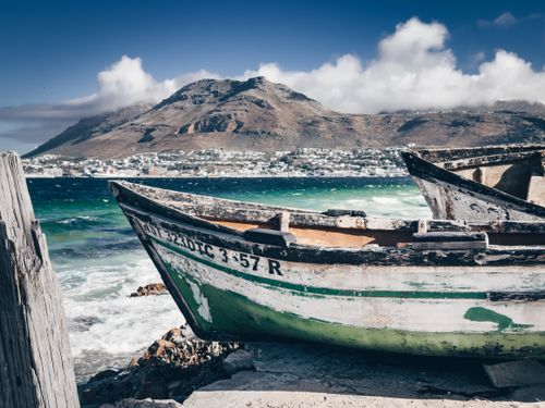 Free A rustic boat rests on the vibrant coast of Cape Town, with mountains in the background, depicting a serene maritime scene. Stock Photo