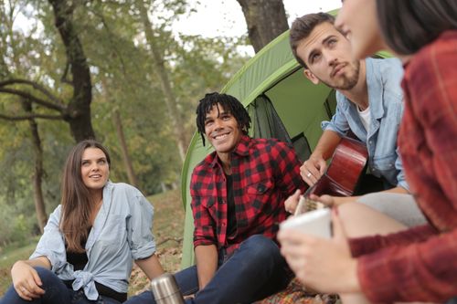 Free Group of young friends in casual clothes sitting on ground singing and playing guitar while spending time together on picnic Stock Photo