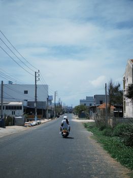 A person rides a motorbike down a quiet urban street under a clear blue sky, showcasing cityscape and lifestyle.