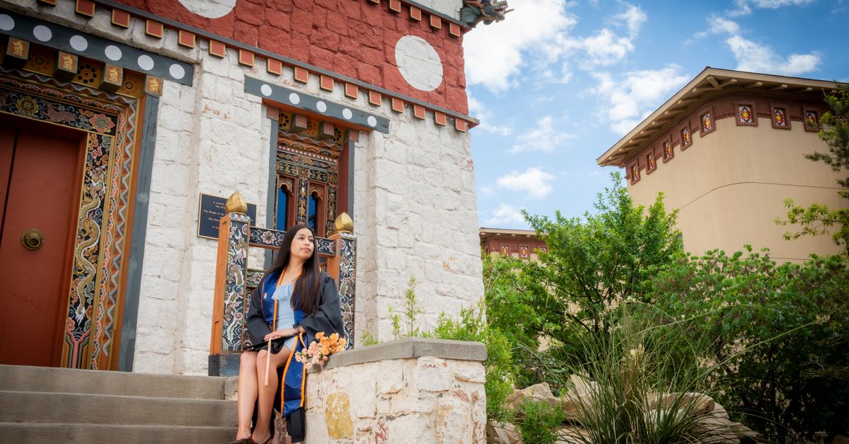 A young woman in a graduation gown sitting on steps outside a colorful, traditional temple building.